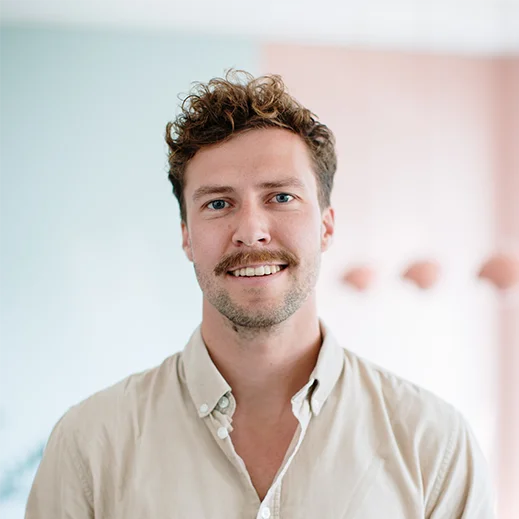 Bild von Benedikt Luber. Teammitglied von TeleClinic. Freundlich blickender junger Mann mit braunen Haaren und braunen Bart innerhalb eines TeleClinic Bürogebäudes.
