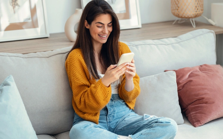 Glückliche Frau sitzt auf dem Sofa und nutzt ihr Smartphone, möglicherweise für eine Online-Arztberatung oder Gesundheits-App.