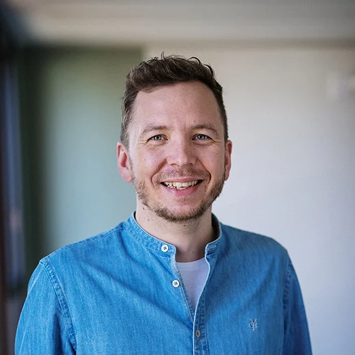 Bild von Matthias Hoyer. Teammitglied von TeleClinic. Freundlich blickender junger Mann mit braunen Haaren und Bart innerhalb eines TeleClinic Bürogebäudes.