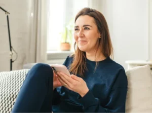 Lächelnde Frau auf Sofa mit Smartphone, symbolisiert Komfort bei digitaler Kommunikation bzw. digitaler Beratung.