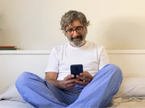 Lächelnder Mann in Pyjama sitzt im Bett und nutzt ein Smartphone, symbolisiert Telemedizin von zu Hause.