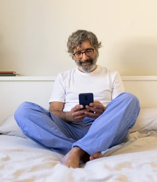 Lächelnder Mann in Pyjama sitzt im Bett und nutzt ein Smartphone, symbolisiert Telemedizin von zu Hause.