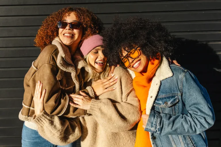 Drei Frauen lachen und umarmen sich, tragen Winterkleidung und Sonnenbrillen.