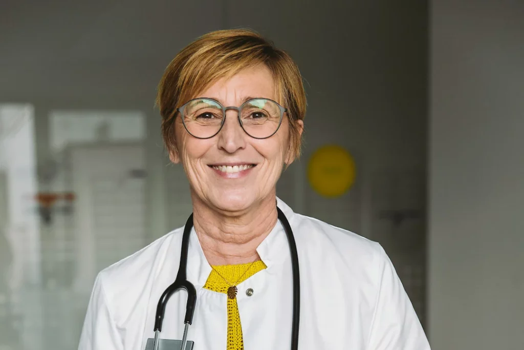 Ärztin mit Brille und Stethoskop lächelt in die Kamera in einer Praxis.